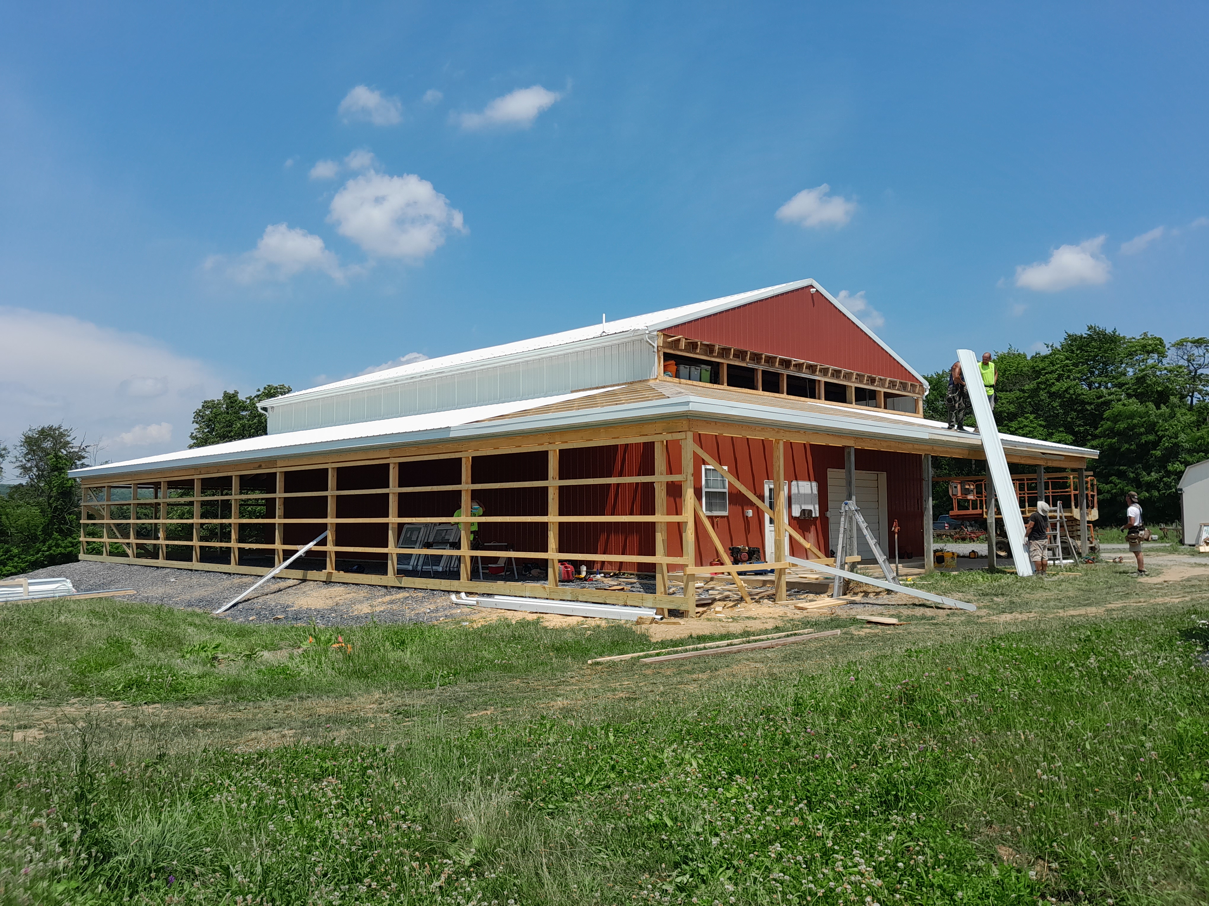Red barn in a field with trees behind. The building has construction around it, with one man on the roof is handed supplies.